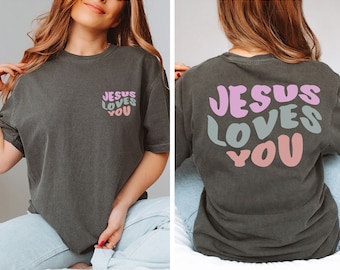 May include: A dark gray t-shirt with the text "Jesus Loves You" in pink, green, and peach on the back. The same text is printed in a smaller size on the front of the shirt.