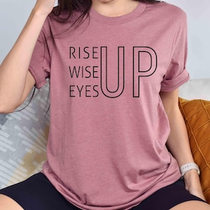 May include: A mauve t-shirt with the text "Rise Up Wise Eyes" printed in black.