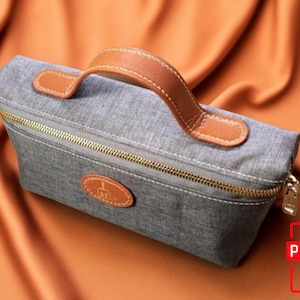 Könnte beinhalten: Eine graue Reißverschlusstasche mit einem braunen Ledergriff. Die Tasche hat einen kleinen Lederfleck mit einem Logo auf der Vorderseite.