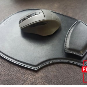 May include: A black leather mouse pad with a built-in wrist rest and a gray wireless mouse. The mouse pad has the word "BIZON" printed on it. The pad is on a brown leather surface.