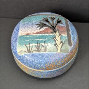 May include: A round ceramic trinket box with a blue and brown glaze. The lid features a painted scene of a palm tree on a beach with waves and mountains in the background.