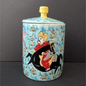 Puede incluir: Un tarro de cerámica con tapa, con un diseño pintado a mano. El tarro es de color azul claro con un patrón floral repetido y una imagen central de una figura sobre un caballo negro. La tapa tiene un pomo amarillo y un diseño floral. El tarro mide unos 13 cm de alto.