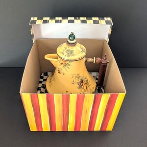 May include: A yellow ceramic teapot with floral designs and a wooden handle sits inside a decorative box. The box has red and yellow stripes on the outside and a black and white checkered pattern inside.