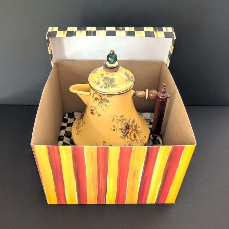 May include: A yellow ceramic teapot with floral designs and a wooden handle sits inside a decorative box. The box has red and yellow stripes on the outside and a black and white checkered pattern inside.