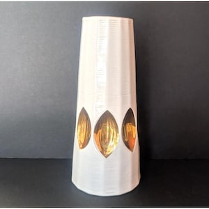 May include: A tall, tapered white ceramic vase featuring a textured, ribbed surface. The exterior is decorated with a horizontal row of three distinct, almond-shaped cutouts that reveal a reflective, metallic gold interior finish.