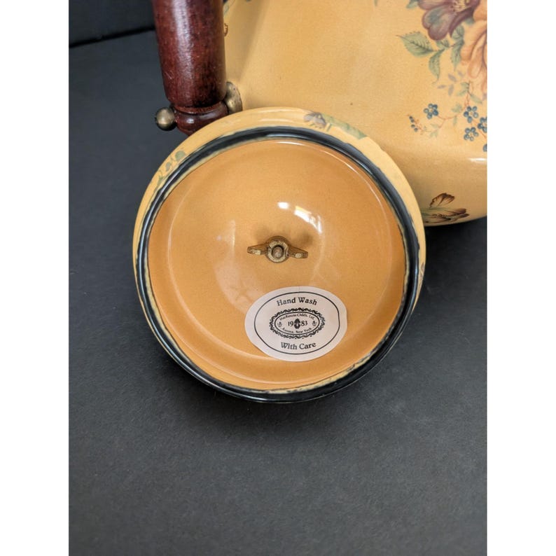 May include: A cream-colored teapot with a floral design and a dark brown handle. The lid is off, revealing a light orange interior with a small metal mechanism. The lid has a black and blue border. The label on the lid reads "Hand Wash With Care."