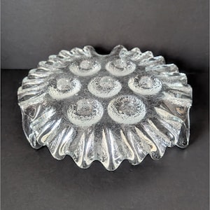 Puede incluir: Un plato de vidrio transparente con un borde festoneado y una superficie texturizada. El plato tiene ocho hendiduras circulares.