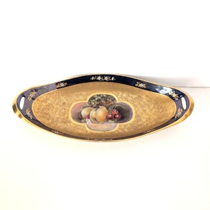 May include: An oval gold-toned serving tray with a dark blue rim and gold handles. The tray features a hand-painted still life of fruit, including peaches and cherries. Delicate floral accents adorn the rim, adding a touch of elegance.