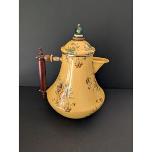 May include: Yellow vintage teapot with floral design. The teapot has a dark brown wooden handle and a lid with a green and gold finial. The teapot is decorated with brown, green, and yellow flowers.