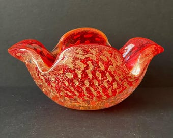 May include: A decorative red glass bowl with a gold speckled pattern. The bowl has a unique shape with flared edges, and is likely used for display or as a decorative accent piece. The bowl is made of glass and has a glossy finish.