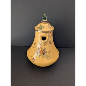 May include: A yellow ceramic teapot with a floral design. The teapot has a lid with a green and gold finial. The body of the teapot is decorated with flowers in shades of brown, purple, and yellow. The spout is rectangular.