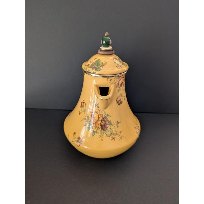 May include: A yellow ceramic teapot with a floral design. The teapot has a lid with a green and gold finial. The body of the teapot is decorated with flowers in shades of brown, purple, and yellow. The spout is rectangular.