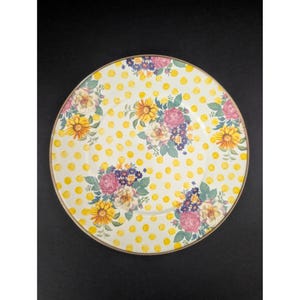 Op de afbeelding: Een ronde, decoratieve plaat met een geel stippenpatroon en bloemboeketten. De boeketten bevatten roze rozen, gele zonnebloemen en paarse bloemen. De plaat heeft een gouden rand en staat tegen een zwarte achtergrond.