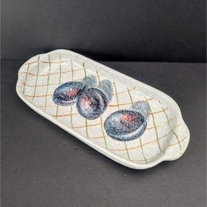 May include: A rectangular ceramic serving tray with a white background and a grid pattern in orange and green. The tray features a hand-painted design of three dark blue plums with red centers and green leaves.