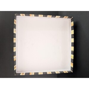 May include: An open, square box with a white interior and a black and tan striped border. The box is empty and sits on a black surface. The stripes are vertical and alternate between black and tan.