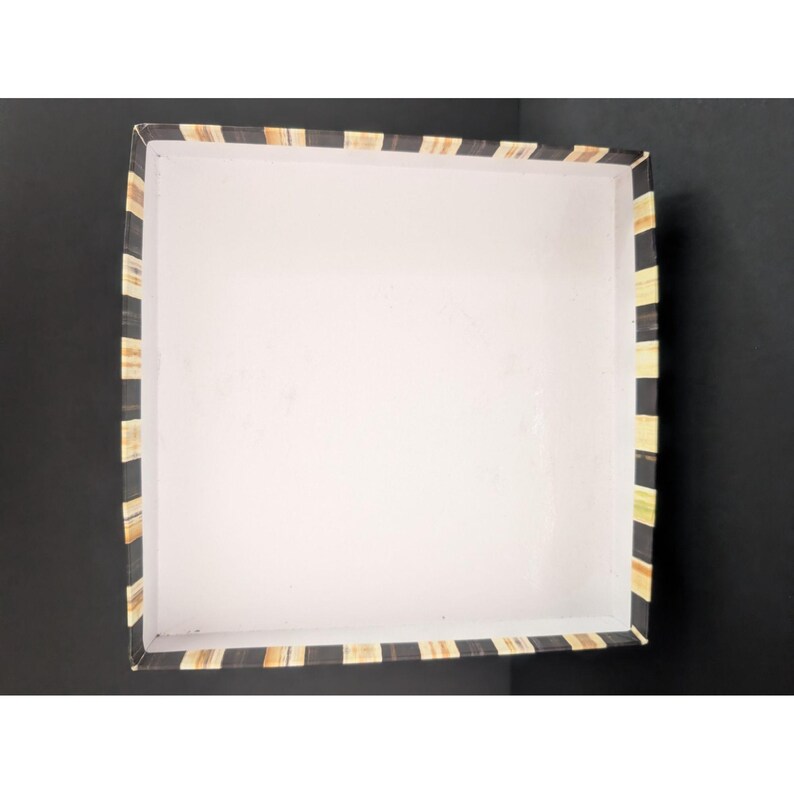 May include: An open, square box with a white interior and a black and tan striped border. The box is empty and sits on a black surface. The stripes are vertical and alternate between black and tan.