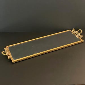 May include: A rectangular gold-toned mirror tray with decorative floral and bow accents on each end. The tray has a mirrored surface and a patterned border. The handles are a twisted rope design.