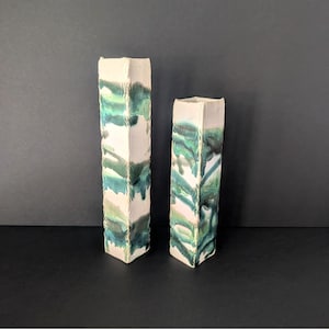 Puede incluir: Dos jarrones rectangulares de cerámica. Los jarrones son blancos con un esmalte verde y azul que gotea por los lados. Los jarrones son de diferentes alturas, el más alto a la izquierda. Los jarrones están sobre una superficie negra.