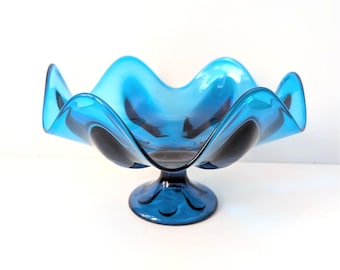 Könnte beinhalten: Eine leuchtend blaue Glasschale mit gewelltem Rand, die auf einem Sockel steht. Die wellenförmigen Kanten der Schale und das dekorative Design des Sockels tragen zu ihrer eleganten Ästhetik bei. Dieses dekorative Stück eignet sich ideal zur Präsentation von Obst oder als Mittelstück.