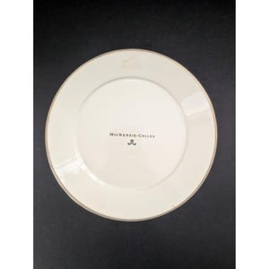 Op de afbeelding: Cr&egrave;mekleurige keramische plaat met een gouden rand. De plaat heeft de tekst "McKenzie-Childs" in zwart, en een klein embleem. De plaat is rond en lijkt ongebruikt.