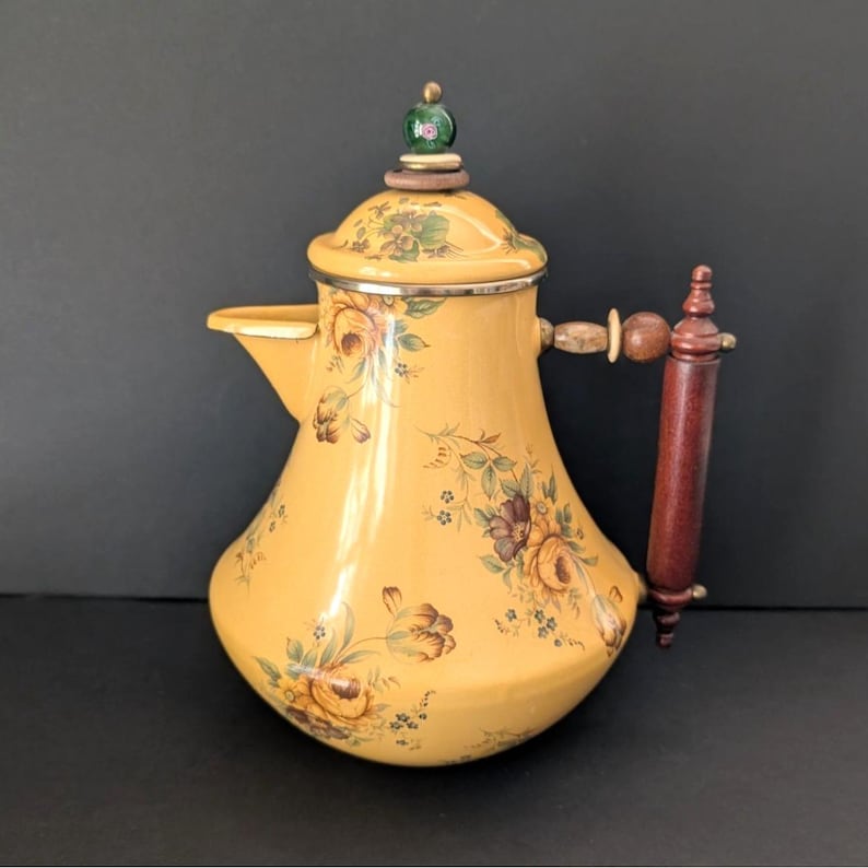 May include: Yellow enamel coffee pot with floral design. The pot has a wooden handle and a decorative top with a green and white accent. The pot is approximately 10 inches tall.