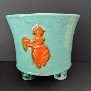 May include: A turquoise ceramic planter with three legs and a raised orange design of a woman in a dress. The planter has a crackled glaze.