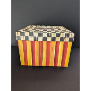 May include: A rectangular decorative box with a black and white checkered lid. The box's sides feature vertical stripes in alternating shades of yellow and red. The box is sitting on a black surface.