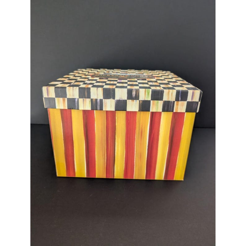 May include: A rectangular decorative box with a black and white checkered lid. The box's sides feature vertical stripes in alternating shades of yellow and red. The box is sitting on a black surface.