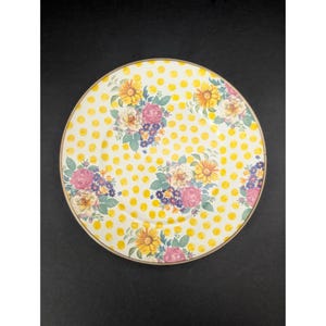 Op de afbeelding: Een ronde, decoratieve plaat met een gele stippenpatroon en bloemstukken. De bloemen zijn in tinten roze, geel en paars, met groene bladeren. De plaat heeft een gouden rand.
