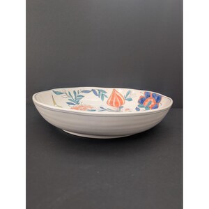 May include: A white ceramic serving bowl with a floral design. The bowl has a wide, shallow shape with a slightly ribbed exterior. The floral pattern includes orange, blue, and green elements. The rim has a thin blue stripe.