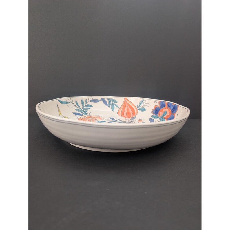 May include: A white ceramic serving bowl with a floral design. The bowl has a wide, shallow shape with a slightly ribbed exterior. The floral pattern includes orange, blue, and green elements. The rim has a thin blue stripe.