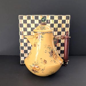 May include: Yellow enamel teapot with floral design and wooden handle. The teapot has a silver rim and a green and white knob on the lid. A wooden stirring stick is next to the teapot. A black and white checkered box is in the background.