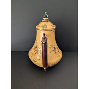 May include: A yellow ceramic teapot with floral designs and a dark brown wooden handle. The lid is topped with a green and gold finial. The teapot has a silver rim and is set against a dark background.