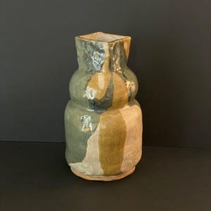 Op de afbeelding: Een handgemaakte keramische vaas met een unieke, organische vorm. De vaas heeft een vierkante opening en is beschilderd met een mix van groene, bruine en bruine kleuren. De vaas is ongeveer 20 cm hoog.