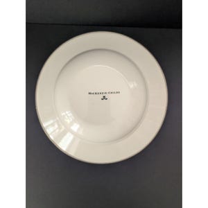 May include: White ceramic plate with a wide rim and a gold-colored edge. The plate has the text "McKENZIE-CHILDS" and a small emblem in the center. The plate is on a dark surface.