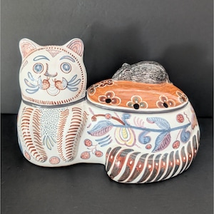 Puede incluir: Una figurita de cerámica de un gato con un ratón durmiendo en su espalda. El gato está pintado en blanco, marrón, azul y amarillo con diseños florales. El ratón está pintado en gris.