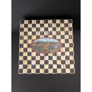 May include: A square box with a black and white checkered pattern. The box has an oval-shaped label in the center with the words "MACKENZIE-CHILDS" and a landscape illustration. The box is set against a black background.