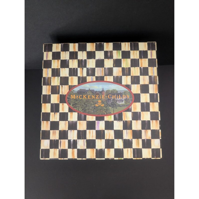 May include: A square box with a black and white checkered pattern. The box has an oval-shaped label in the center with the words "MACKENZIE-CHILDS" and a landscape illustration. The box is set against a black background.
