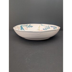 May include: A white ceramic bowl with a floral design. The bowl has a slightly irregular shape and a blue line around the rim. The floral pattern includes blue and green leaves and pink and yellow flowers.