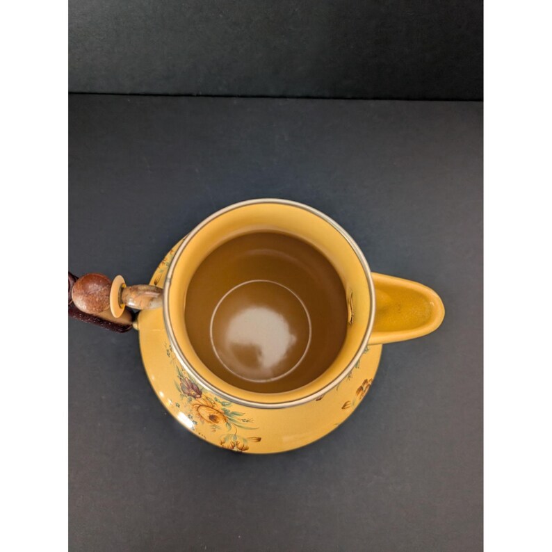 May include: Yellow ceramic teacup and saucer set with floral designs. The teacup has a handle and a brown interior. The saucer is round and has a matching floral pattern. The set is placed on a dark surface.