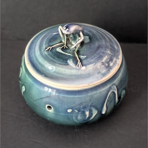 May include: A ceramic jar with a lid, featuring a frog figurine. The jar is a blend of blue and green hues, with a textured surface and decorative patterns. The frog is silver-colored, adding a contrasting element to the design.