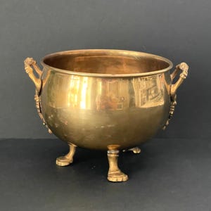 May include: A vintage brass bowl with three small feet and two ornate handles. The bowl has a rounded shape and a polished finish, reflecting the surrounding environment. The handles feature decorative detailing, adding to the antique aesthetic.