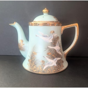 Puede incluir: Una tetera de porcelana azul pálido con una tapa con remate dorado, un asa y un pico dorados. El cuerpo está decorado con aves blancas en vuelo y motivos florales dorados, con una banda dorada que rodea el borde superior de la tetera.