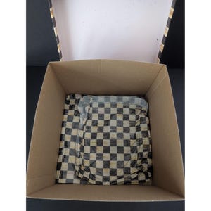 May include: An open cardboard box with a black and white checkered pattern tissue paper inside. The box is beige with black and white stripes on the lid's edge. The tissue paper is folded and crumpled, partially obscuring the contents.