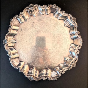 May include: A silver serving tray with a decorative, scalloped edge and an embossed floral design. The tray has a reflective surface and ornate handles. A vintage piece, suitable for serving food or as a decorative item.