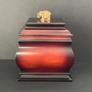 May include: A wooden box with a rich, reddish-brown finish and a small, gold-colored elephant figurine on top. The box has a tiered design with curved sides and a dark base.