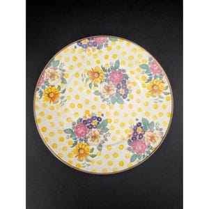 Op de afbeelding: Een decoratieve plaat met een witte achtergrond en gele stippen. De plaat heeft bloemstukken van roze rozen, gele zonnebloemen en blauwe bloemen. De rand van de plaat heeft een gouden rand.