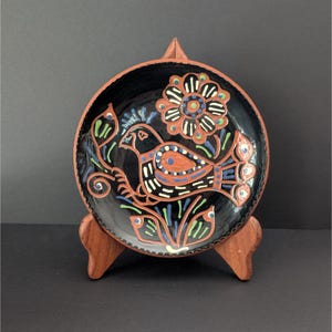 May include: A black ceramic plate with a hand-painted bird and floral design. The bird features orange, blue, and white details. The plate is displayed on a wooden stand, showcasing the folk art style.