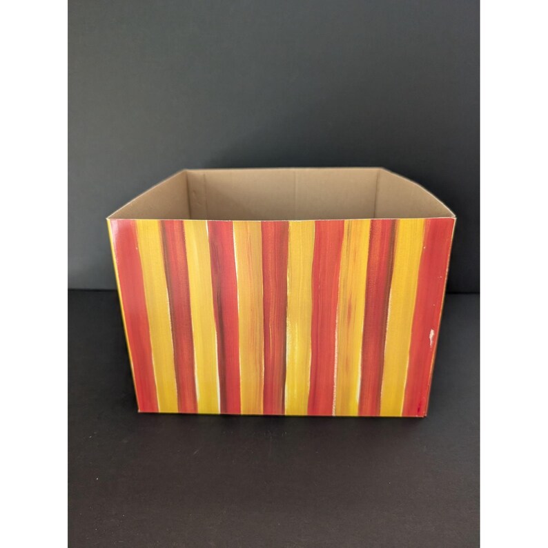 May include: A rectangular cardboard box with a hand-painted design of vertical red and yellow stripes. The box has a natural cardboard interior and is set against a dark background.