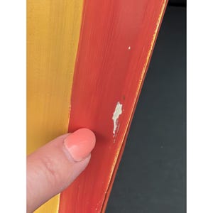 May include: Close-up of a section of a painted wooden object. The object is divided into two sections, one yellow and one red. The red section has some paint chipping. A finger with pink nail polish is touching the object.